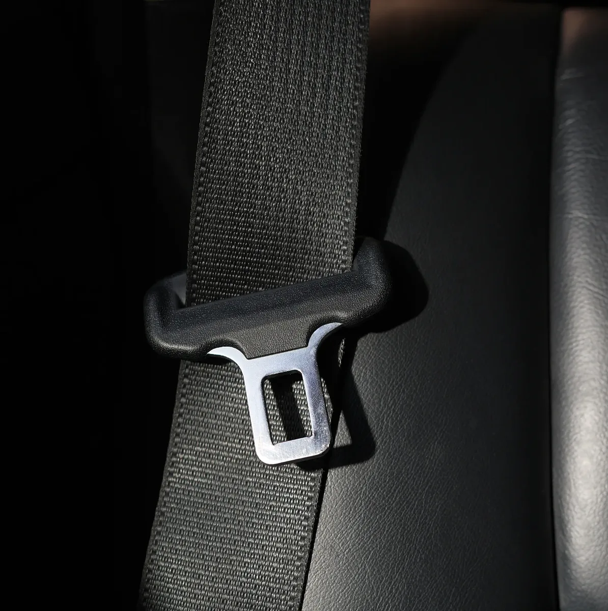 Black seat belt webbing and polished metal tongue plate photographed against a dark leather seat